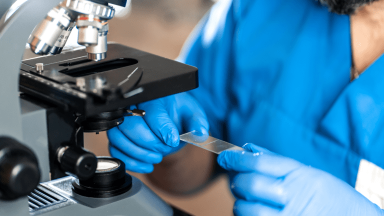 Pesquisador em laboratório manuseando lâmina de análise em microscópio, representando chamada da FAPESP e NWO para materiais avançados em saúde.
