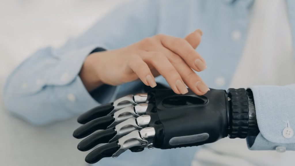 Mão humana tocando uma prótese robótica, simbolizando tecnologia assistiva, acessibilidade e inclusão por meio da inovação