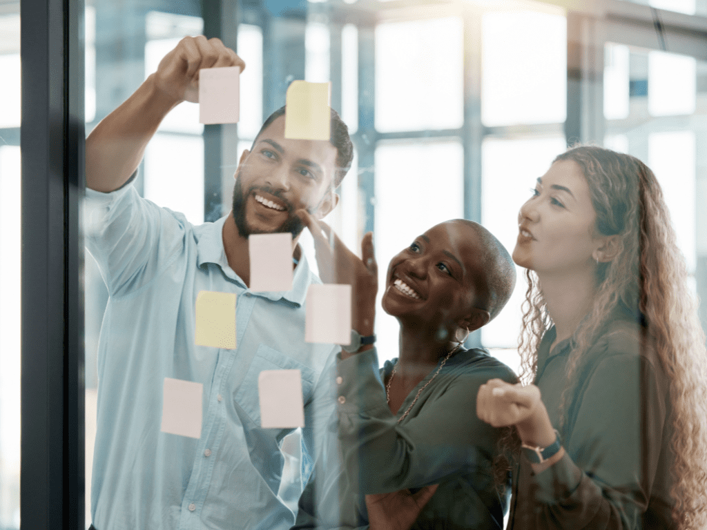 Grupo de profissionais sorrindo e organizando post-its em parede de vidro, representando recrutamento, gestão de talentos e trabalho colaborativo.