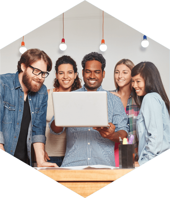 Grupo diverso de profissionais sorrindo enquanto colaboram diante de um laptop em ambiente criativo com luminárias pendentes.