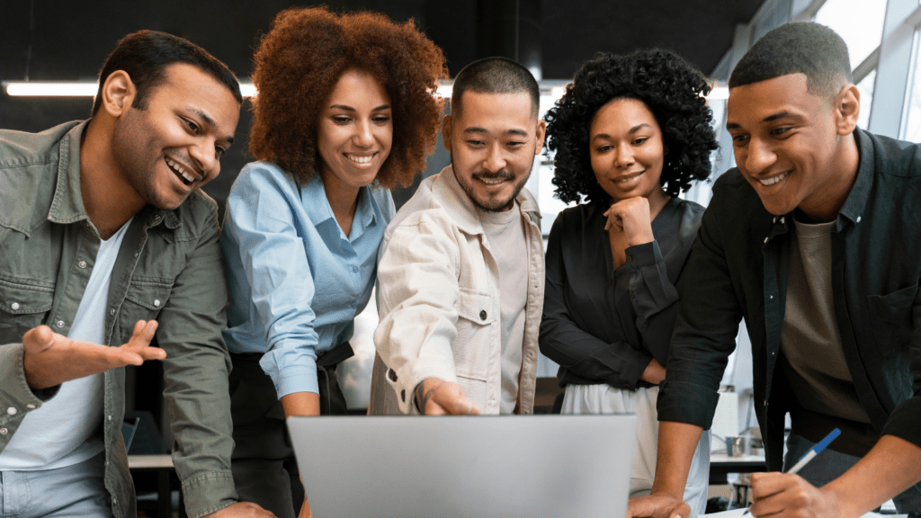Cinco profissionais reunidos em volta de um notebook, um deles apontando para a tela; grupo diverso sorrindo e colaborando em um escritório