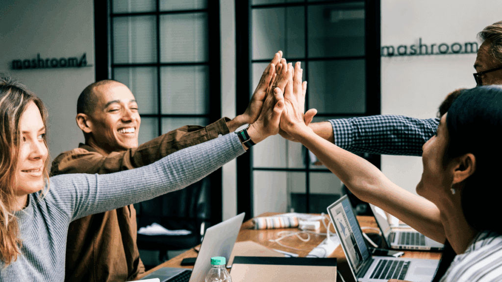 Equipe em escritório celebrando com high-five sobre a mesa com notebooks, simbolizando parceria na captação de recursos para inovação.